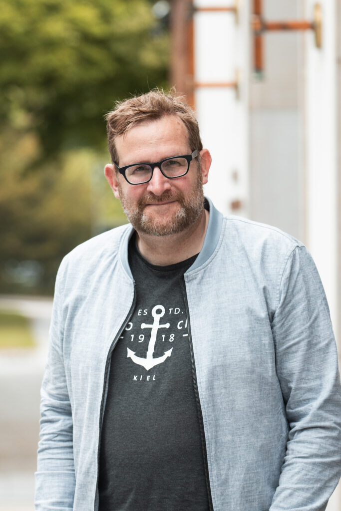 Der Abgeordnete Jan Kürschner steht in einer grauen Jacke mit Reißverschluss und einem dunkelgrauen T-Shirt mit einem Anker und der Aufschrift "Kiel" darunter vor dem Kieler Landtag.