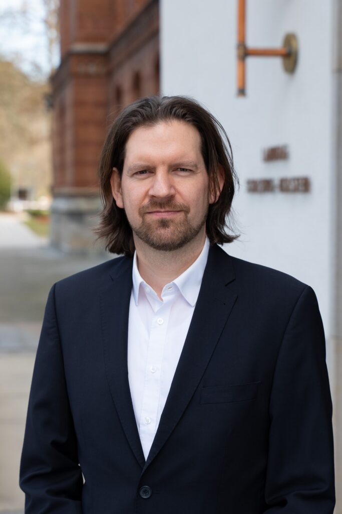 Abgebildet ist der Abgeordnete Sebastian Bonau. Er steht im Anzug vor dem Eingang des Landtages.