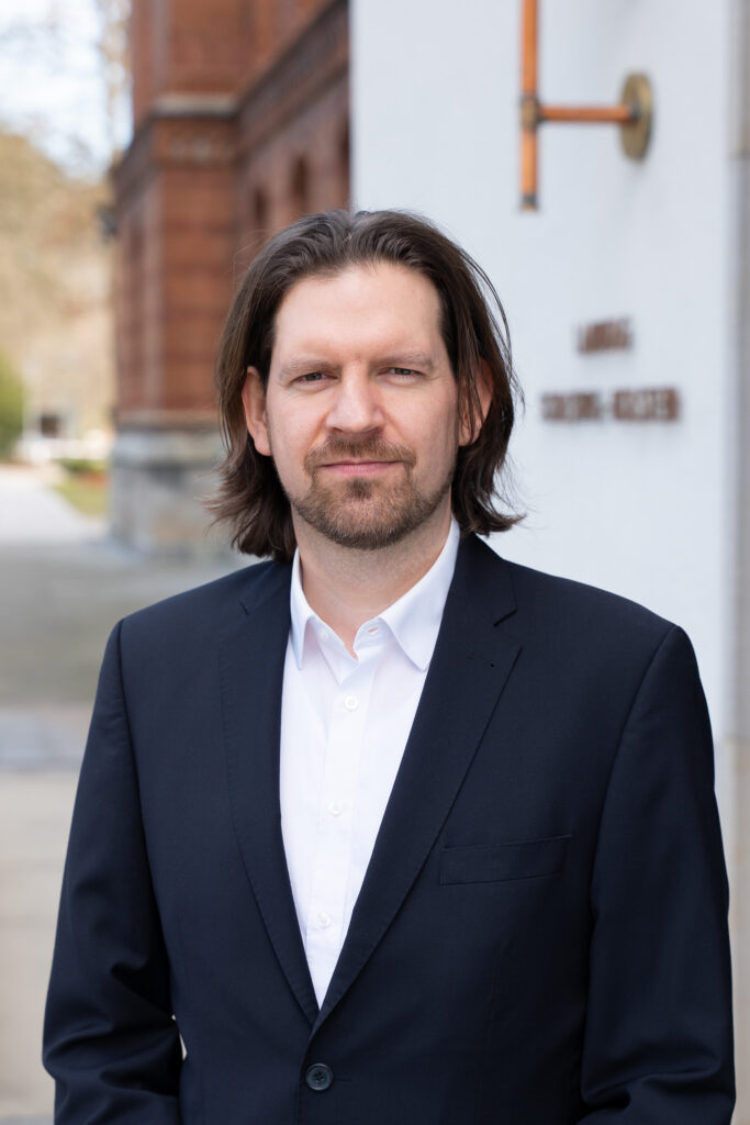 Abgebildet ist der Abgeordnete Sebastian Bonau. Er steht im Anzug vor dem Eingang des Landtages.